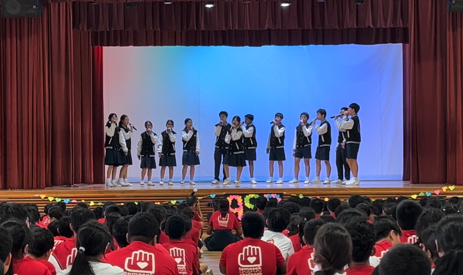 Students in matching jackets and uniforms sing on a stage before a large seated audience in red shirts.